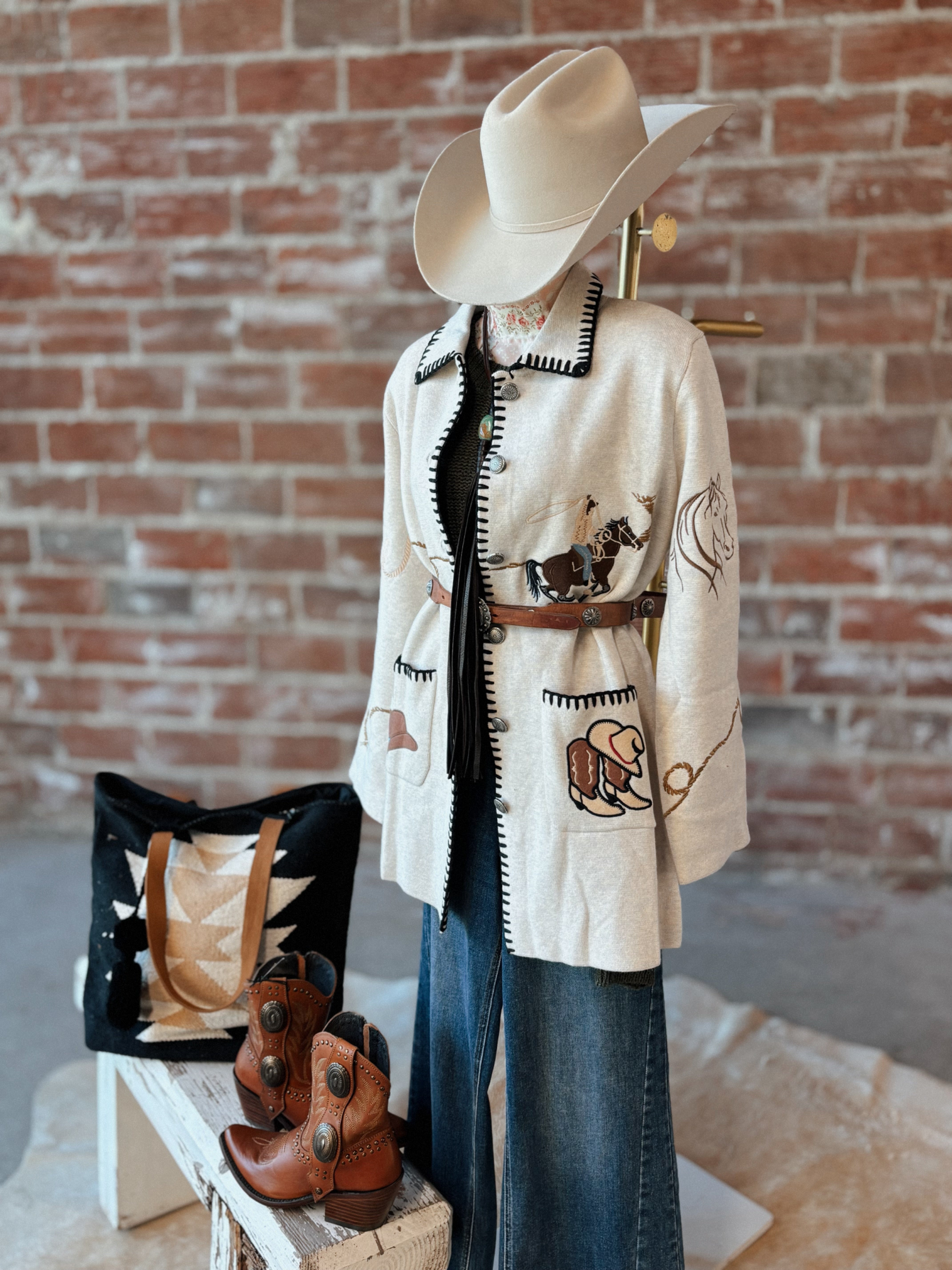 Mannequin wearing a cowboy outfit with a hat, jacket, boots, and bag against a brick wall.