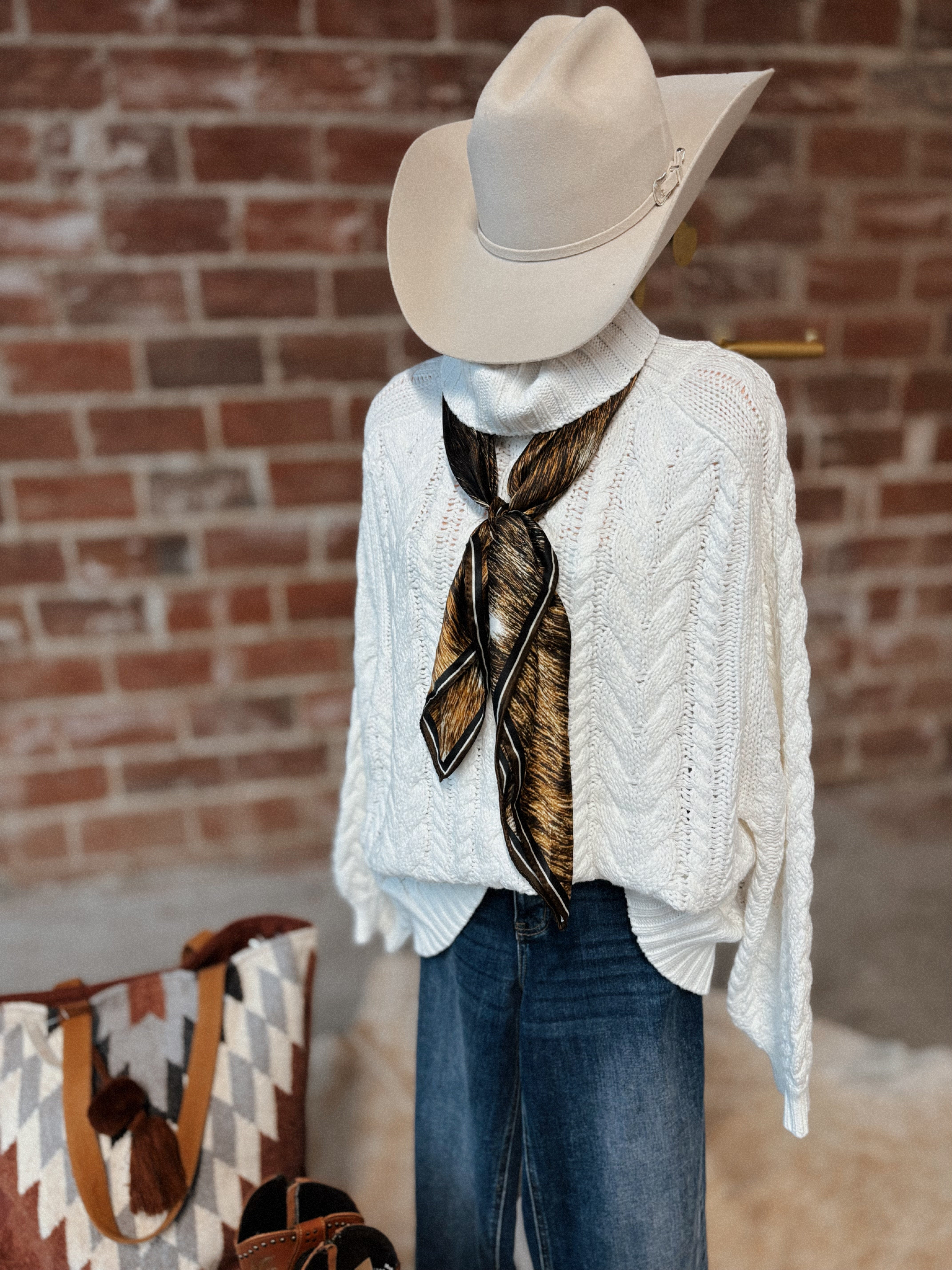 Person wearing a white cable knit sweater, blue jeans, and a beige cowboy hat with a patterned scarf, standing against a brick wall.
