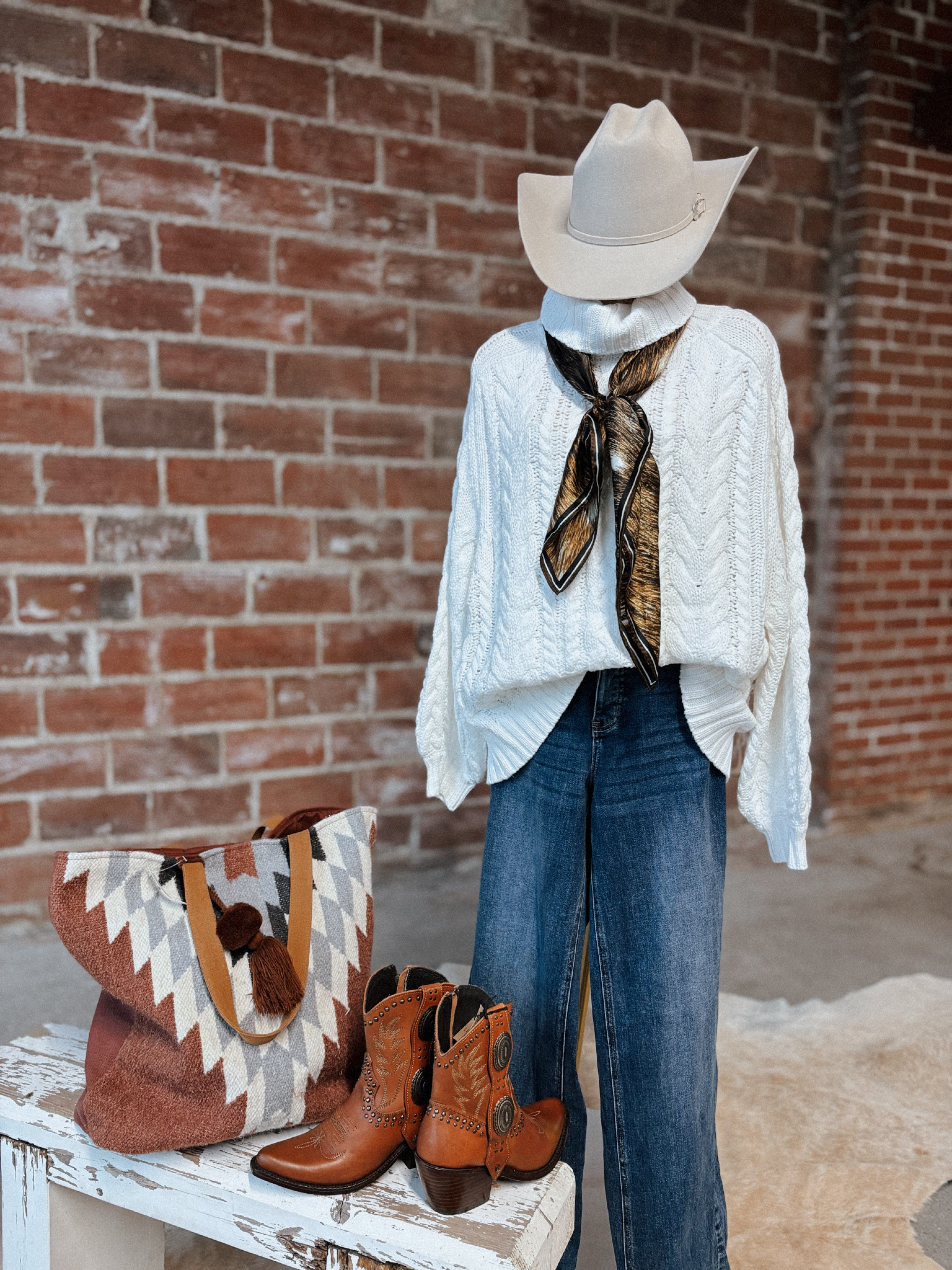 Mannequin wearing a white sweater, blue jeans, and cowboy hat with a scarf, next to brown boots and a patterned bag against a brick wall.