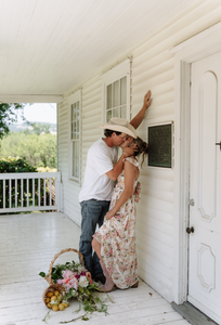 Couple embracing on a white porch with a basket of flowers and a plaque on the wall.
